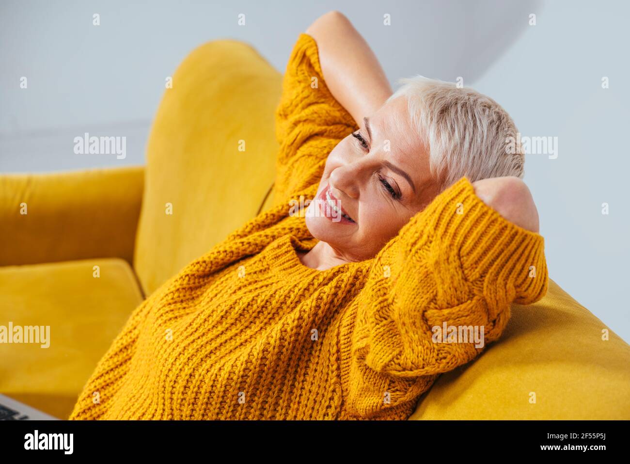Smiling woman with hands behind back looking away while resting on sofa Stock Photo