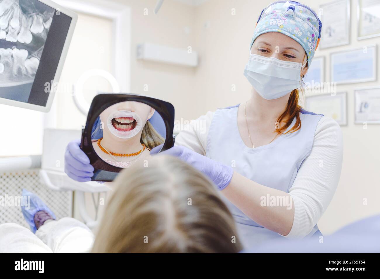 Female dentists holding mirror reflecting mouth of little girl wearing