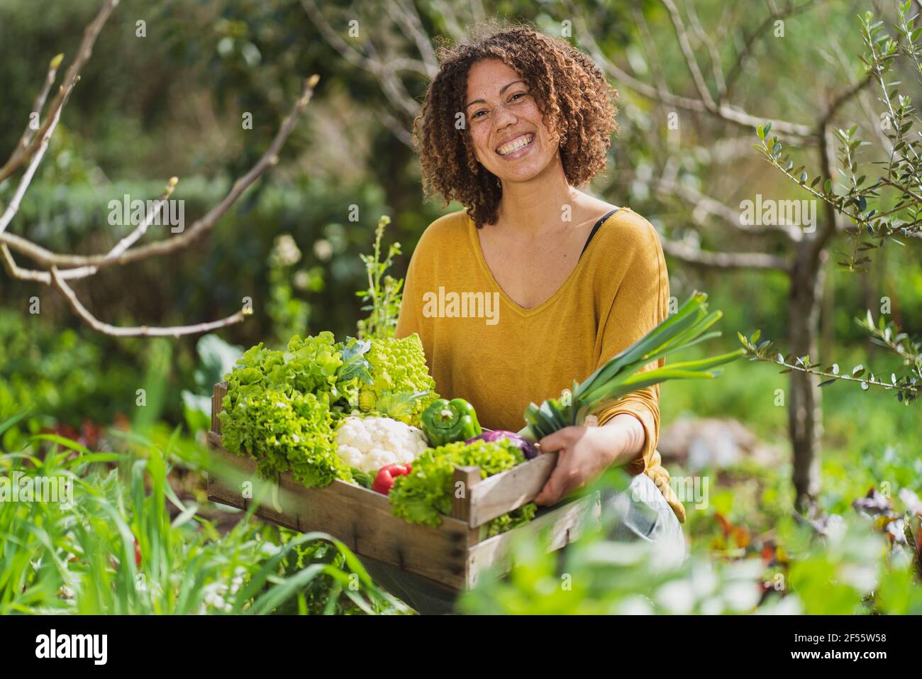 Smiling woman holding a crate with freshly picked organic vegetables in permaculture garden Stock Photo