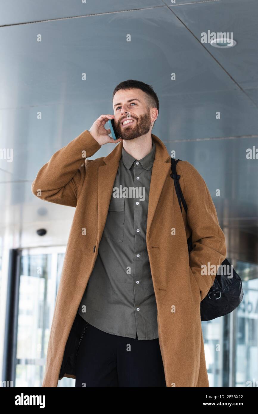 Male entrepreneur talking on mobile phone while standing with hands in pockets outdoors Stock Photo