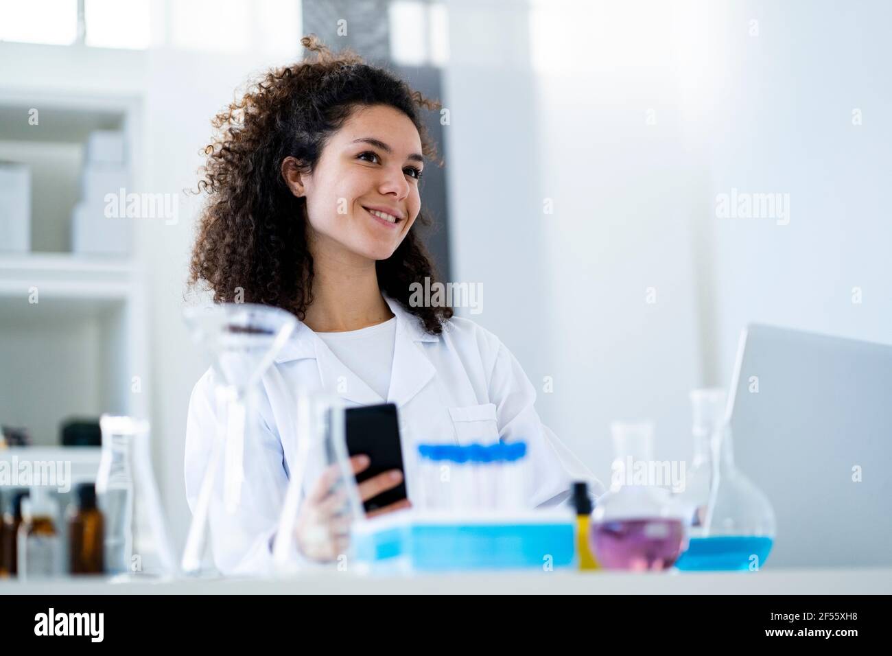 Smiling female scientist with smart phone looking away in hospital Stock Photo