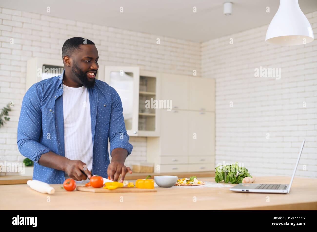 Concentrated african man cooking while watching a video lesson on a laptop, standing at the table top with ingredients for the dish, on the modern kitchen, using knife to cut vegetables on board Stock Photo