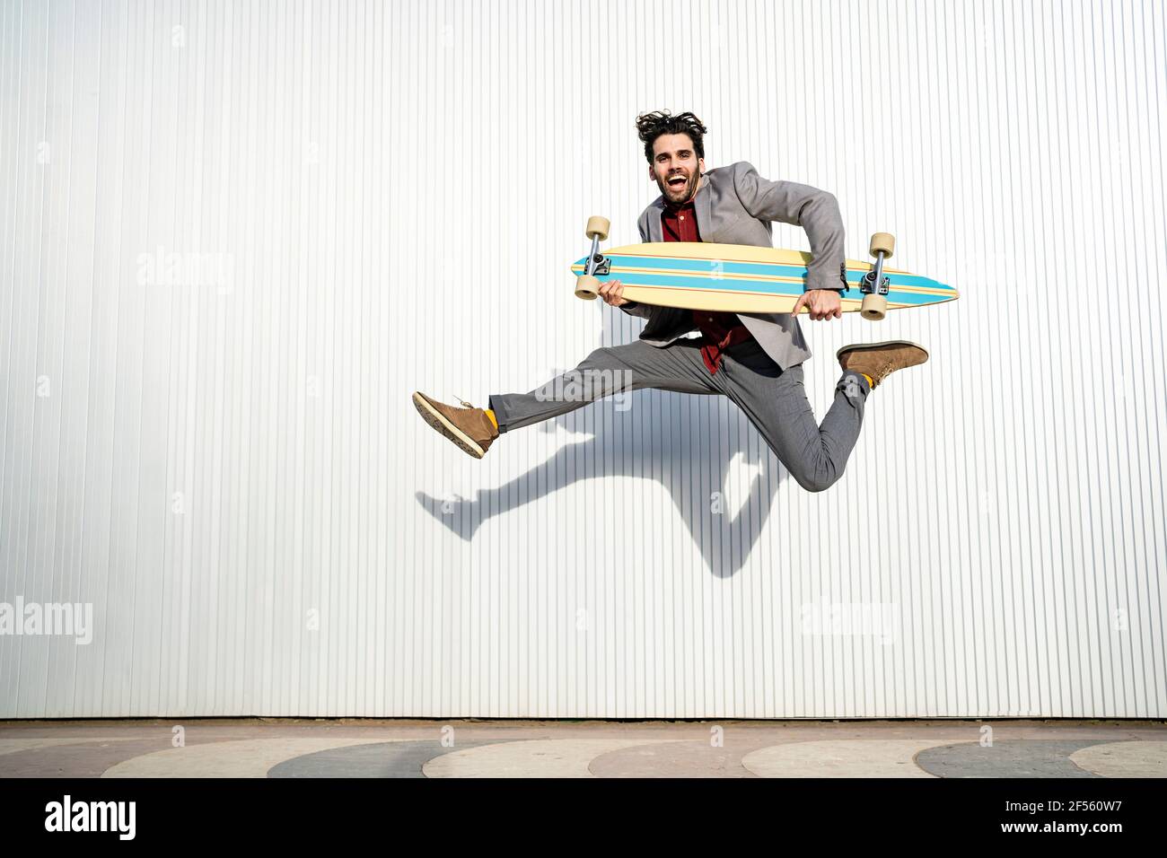 Cheerful man holding longboard while jumping high in front of wall Stock Photo