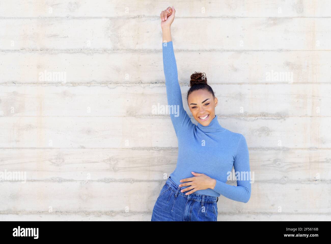 Young woman smiling while standing with hand raised against wall Stock Photo