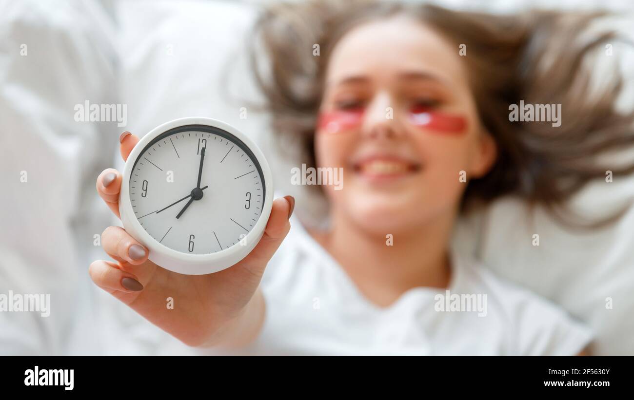 Alarm clock in female hand. Happy young woman wake up after sleeping in bed holding side alarm clock. Morning beauty routine. Woman use eye patches Stock Photo