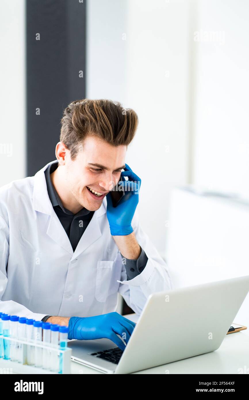 Smiling scientist talking on smart phone while using laptop in laboratory Stock Photo