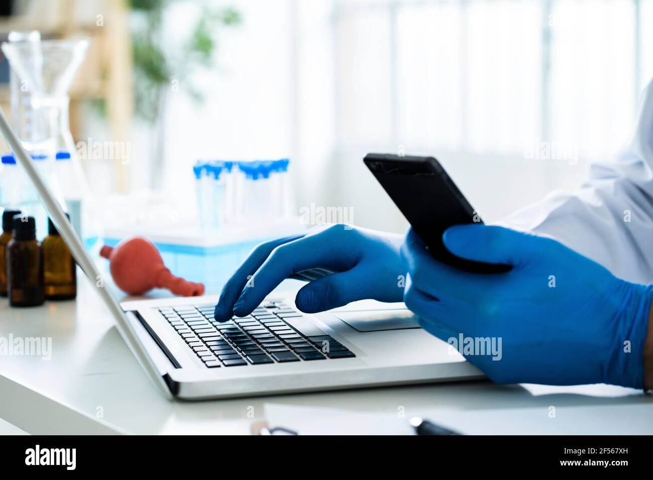 Scientist holding smart phone while using laptop at desk in laboratory Stock Photo