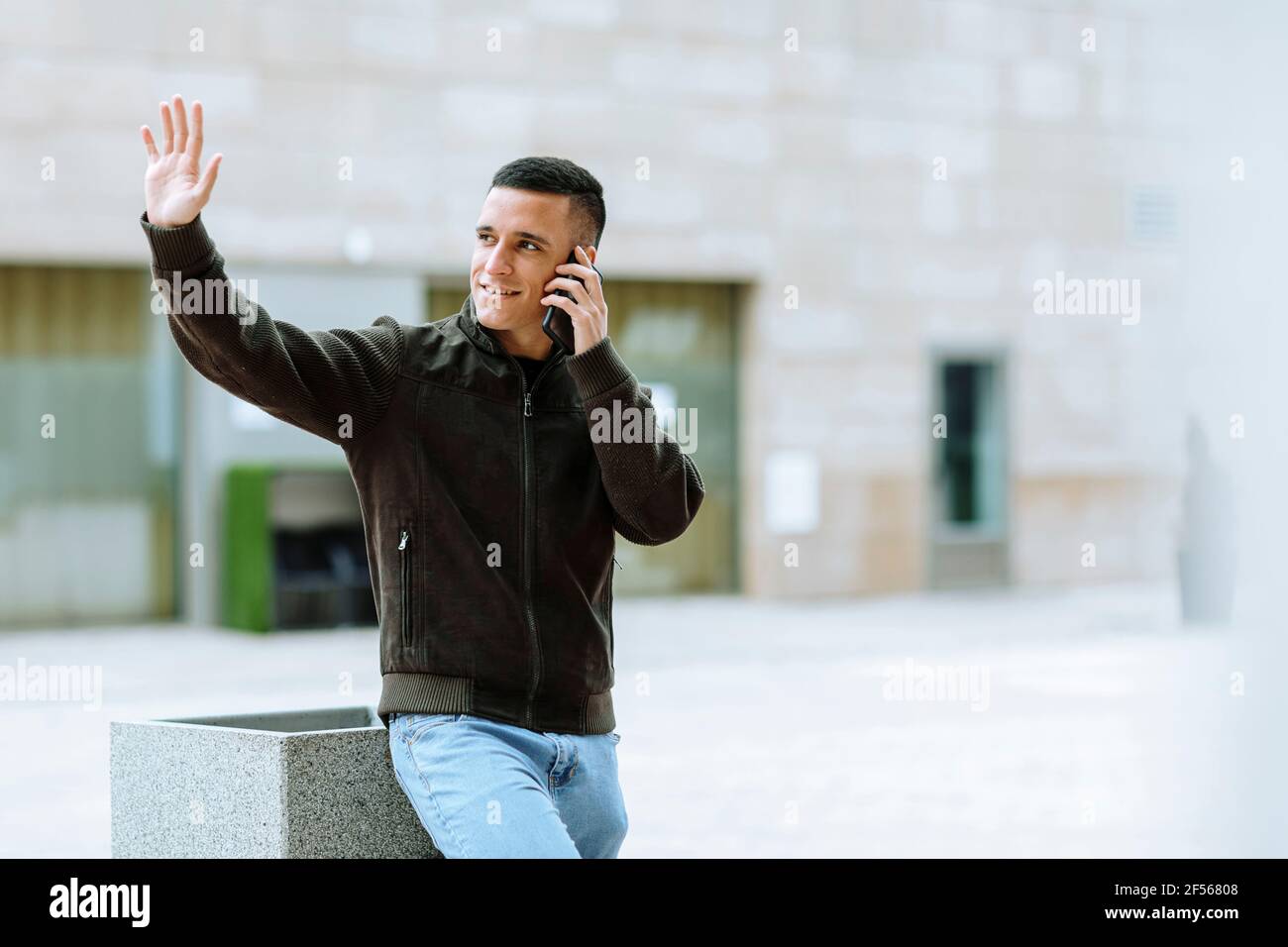 Smiling man waving while talking on mobile phone against building Stock Photo