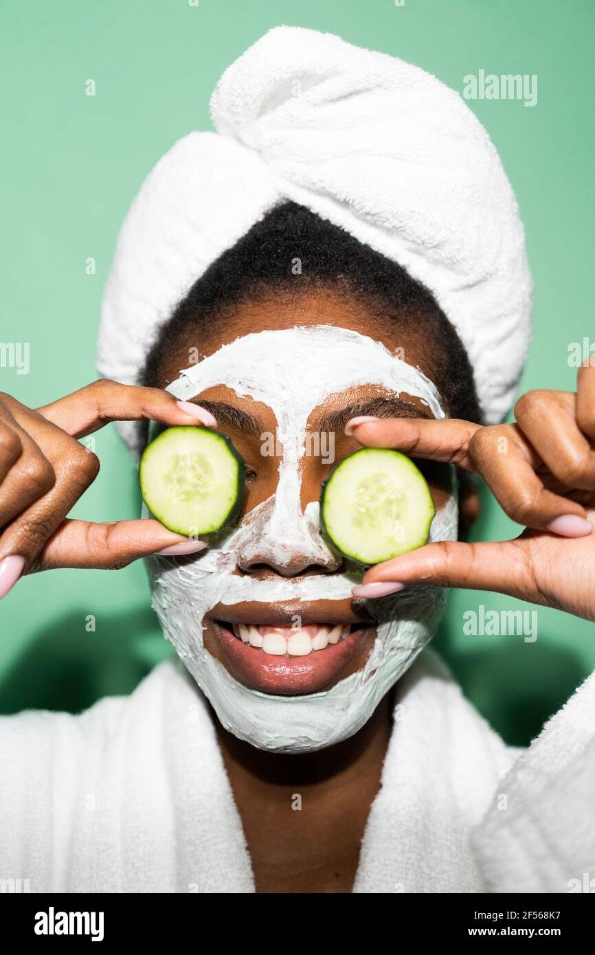 Smiling woman with facial mask covering eyes with cucumber while standing against green background Stock Photo