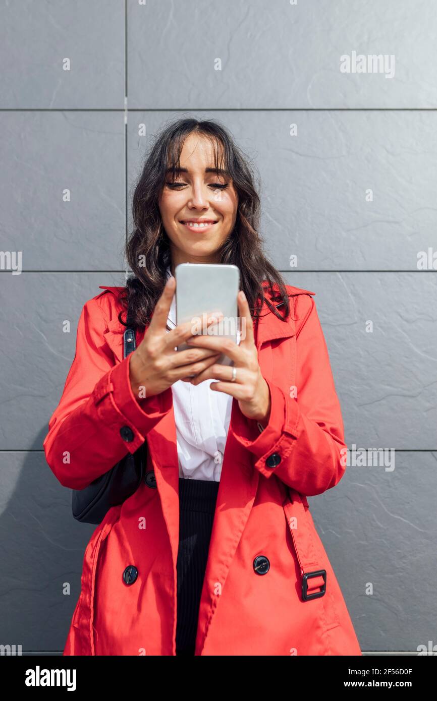 Smiling young businesswoman using phone while standing against gray color wall Stock Photo