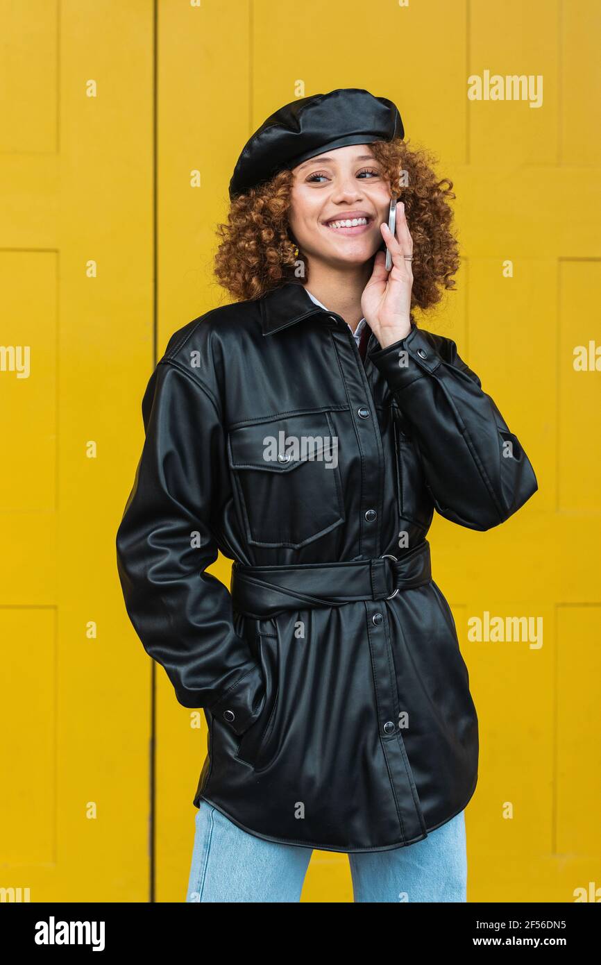 Fashionable teenage girl talking on mobile phone while standing with hands in pockets against door Stock Photo