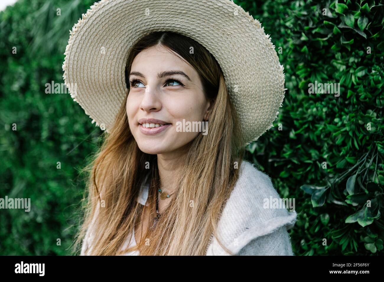 Thoughtful young woman in hat by green plant wall Stock Photo