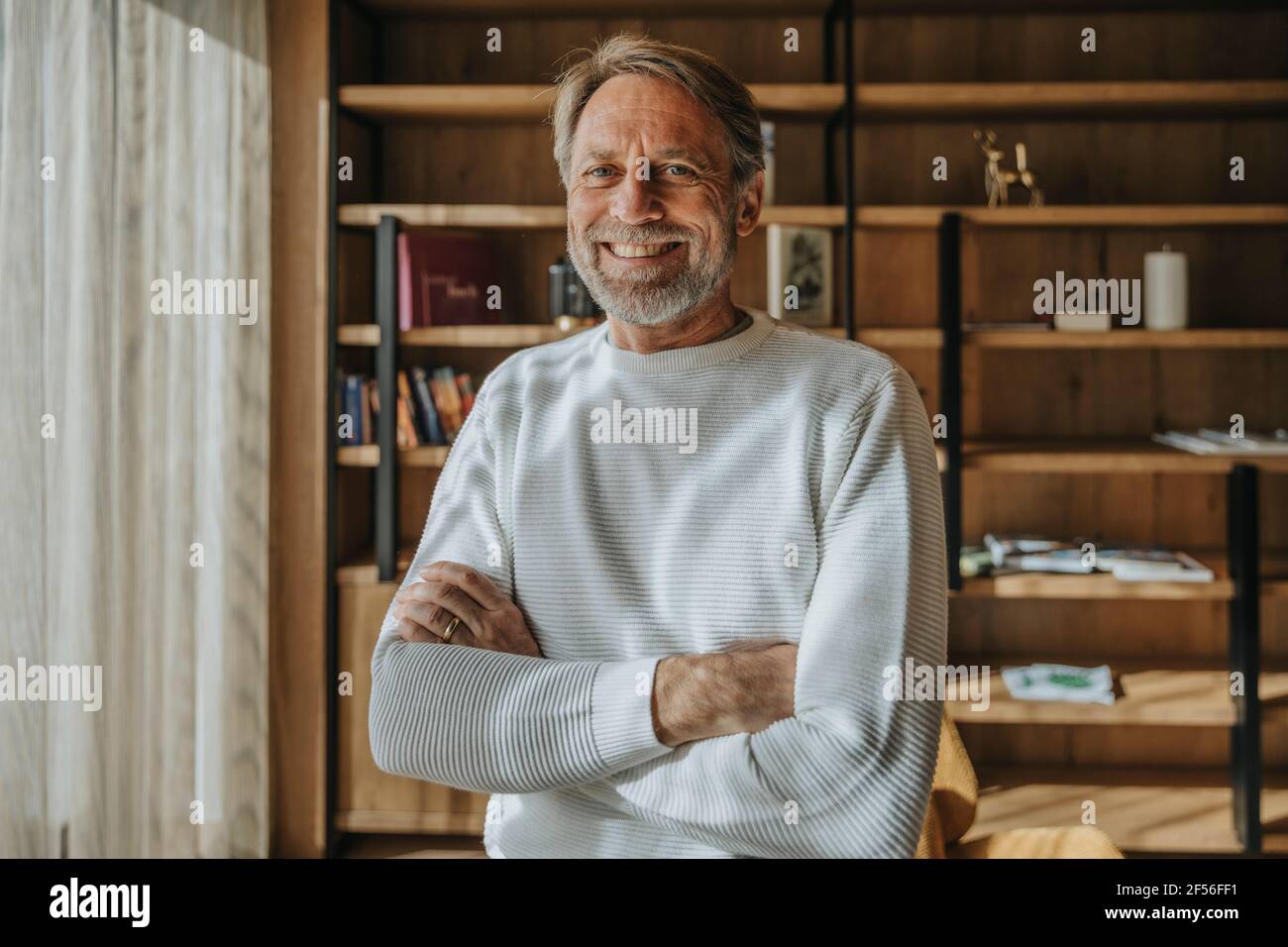 Smiling mature man with arms crossed standing against shelf at home ...