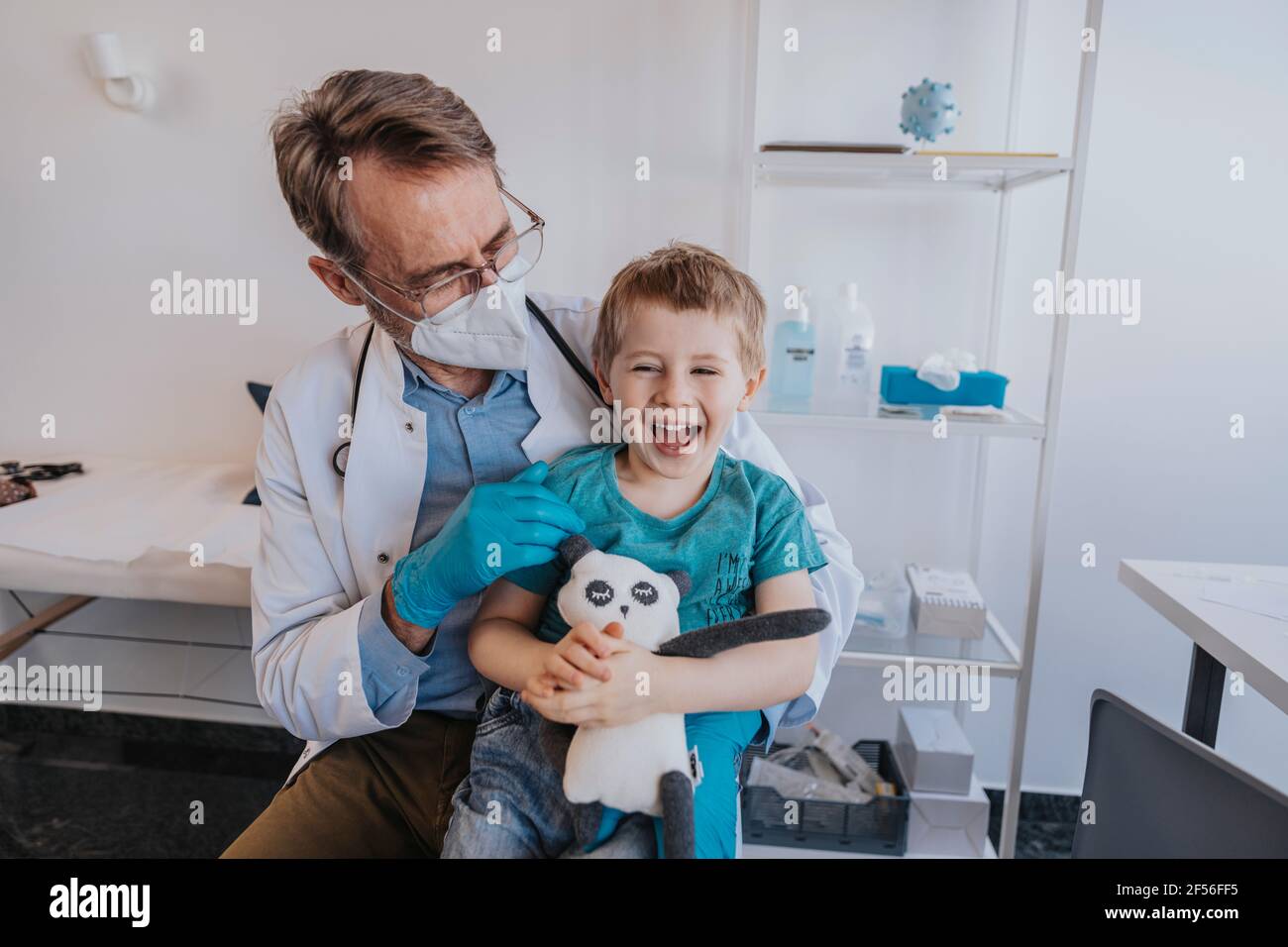 Male pediatrician looking at cheerful boy with toy while sitting at medical examination room Stock Photo