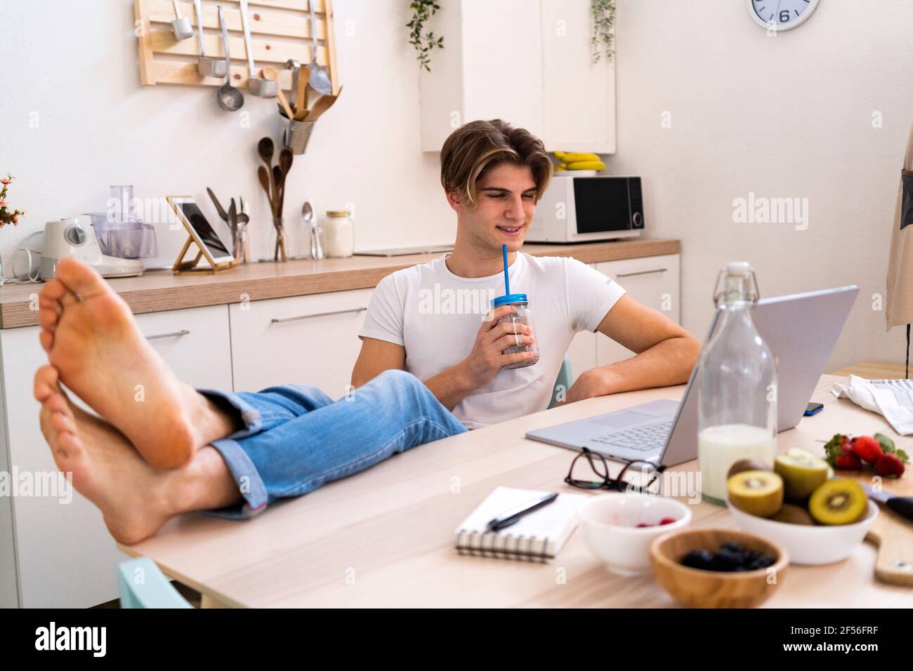 Relaxed young man drinking smoothie while sitting with laptop at table in kitchen Stock Photo