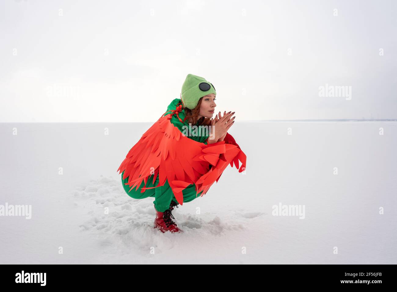 Woman in squatting position while wearing wings with green costume on snow by sky Stock Photo