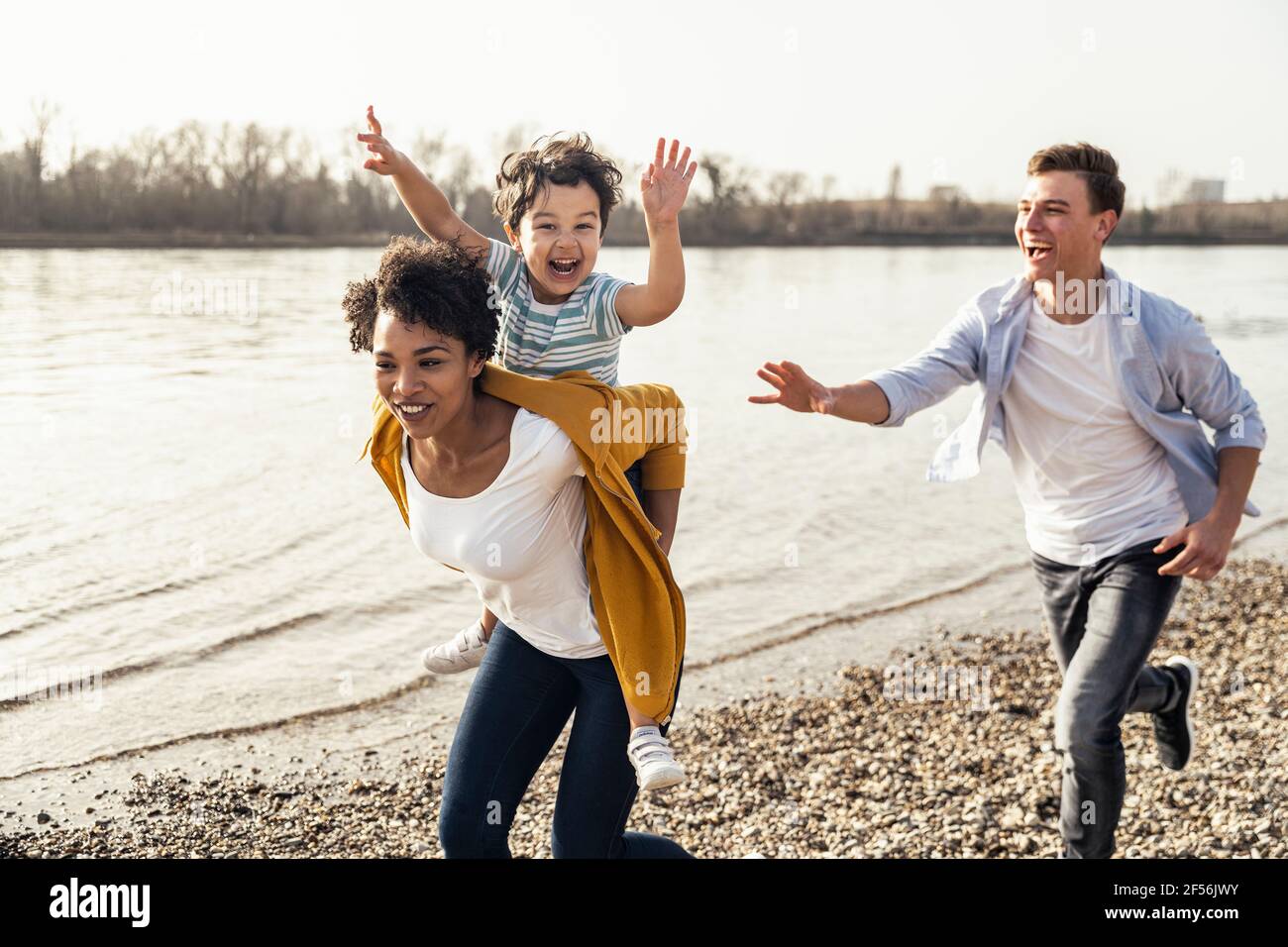 Father running behind mother piggybacking son while running by lakeshore Stock Photo