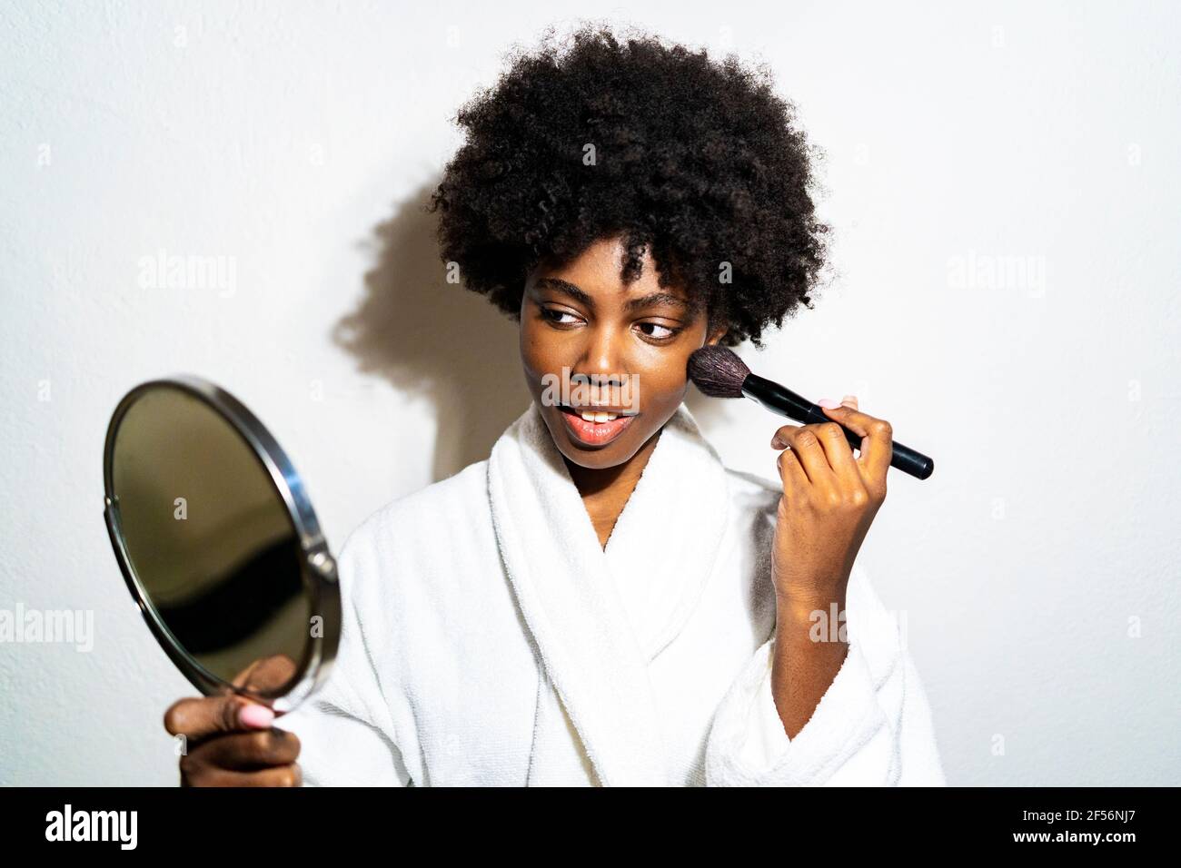 Curly hair woman in bathrobe doing makeup while standing against white