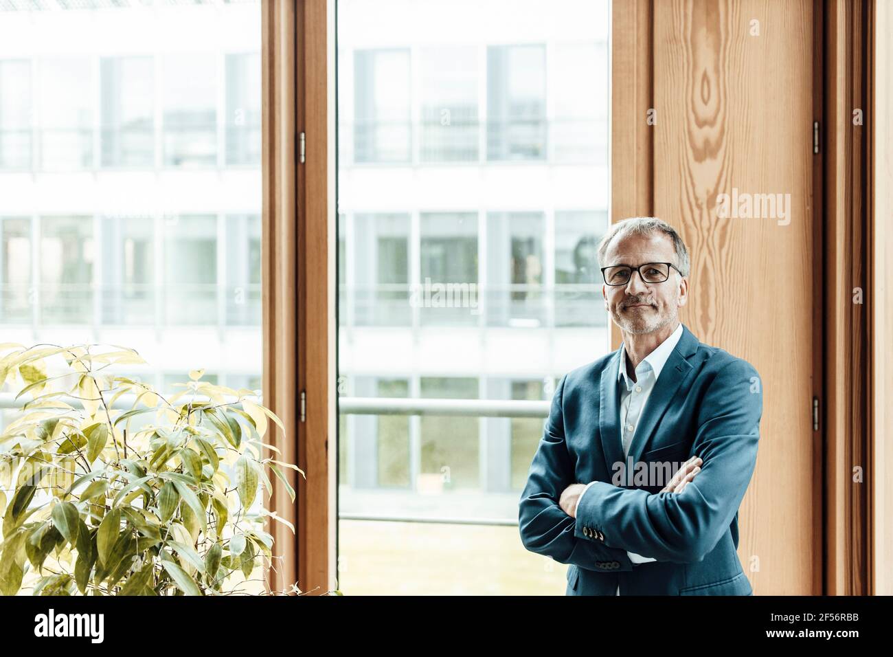 Confident male entrepreneur with arms crossed standing at glass window ...