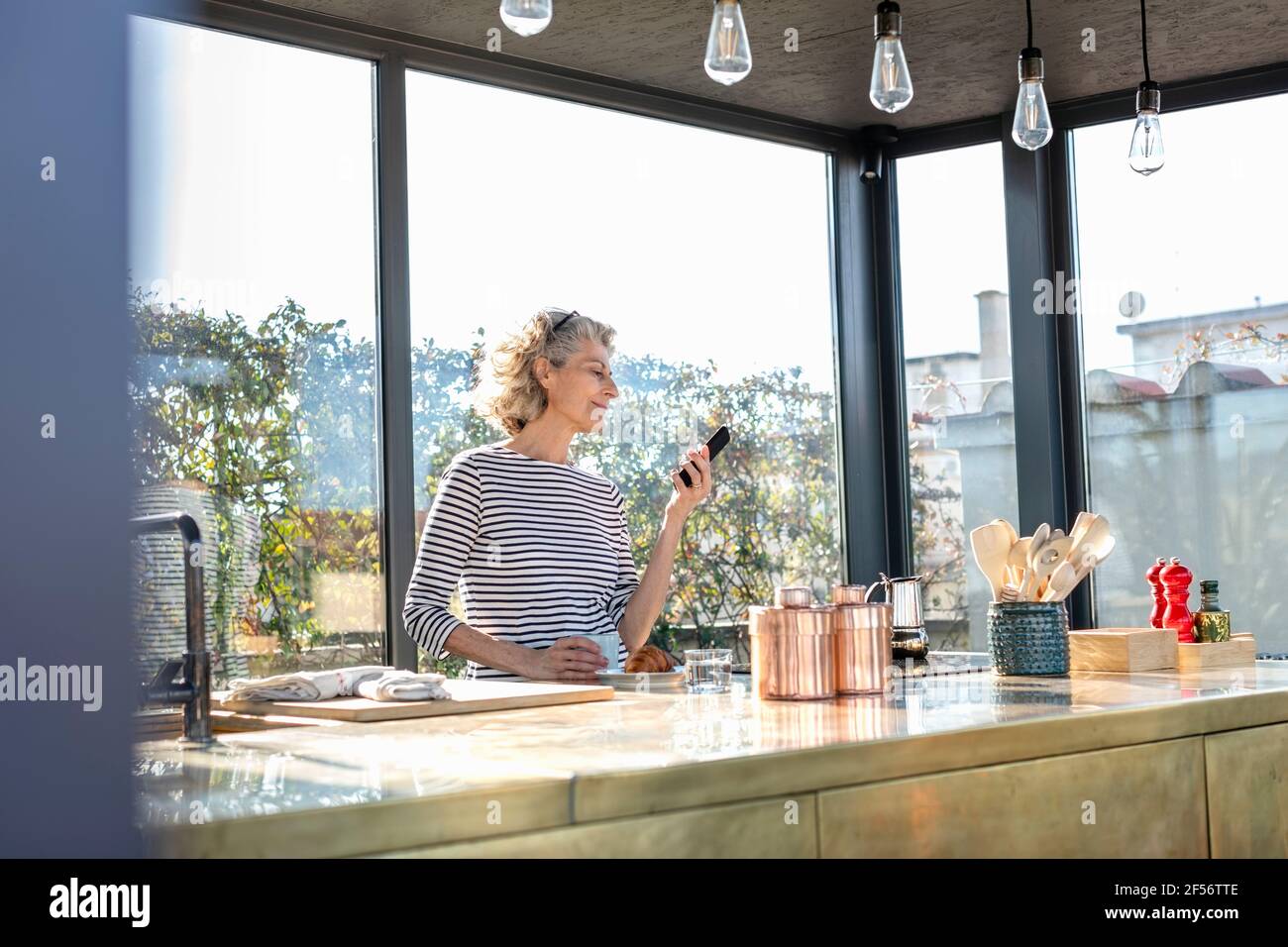 Woman using mobile phone while standing by kitchen island at home Stock Photo