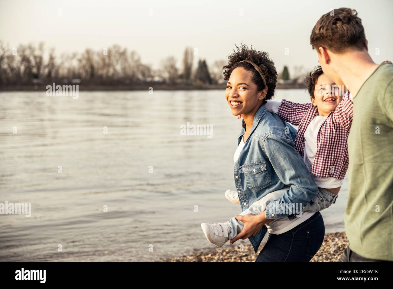 Smiling mother piggybacking son while walking with father by lake Stock Photo