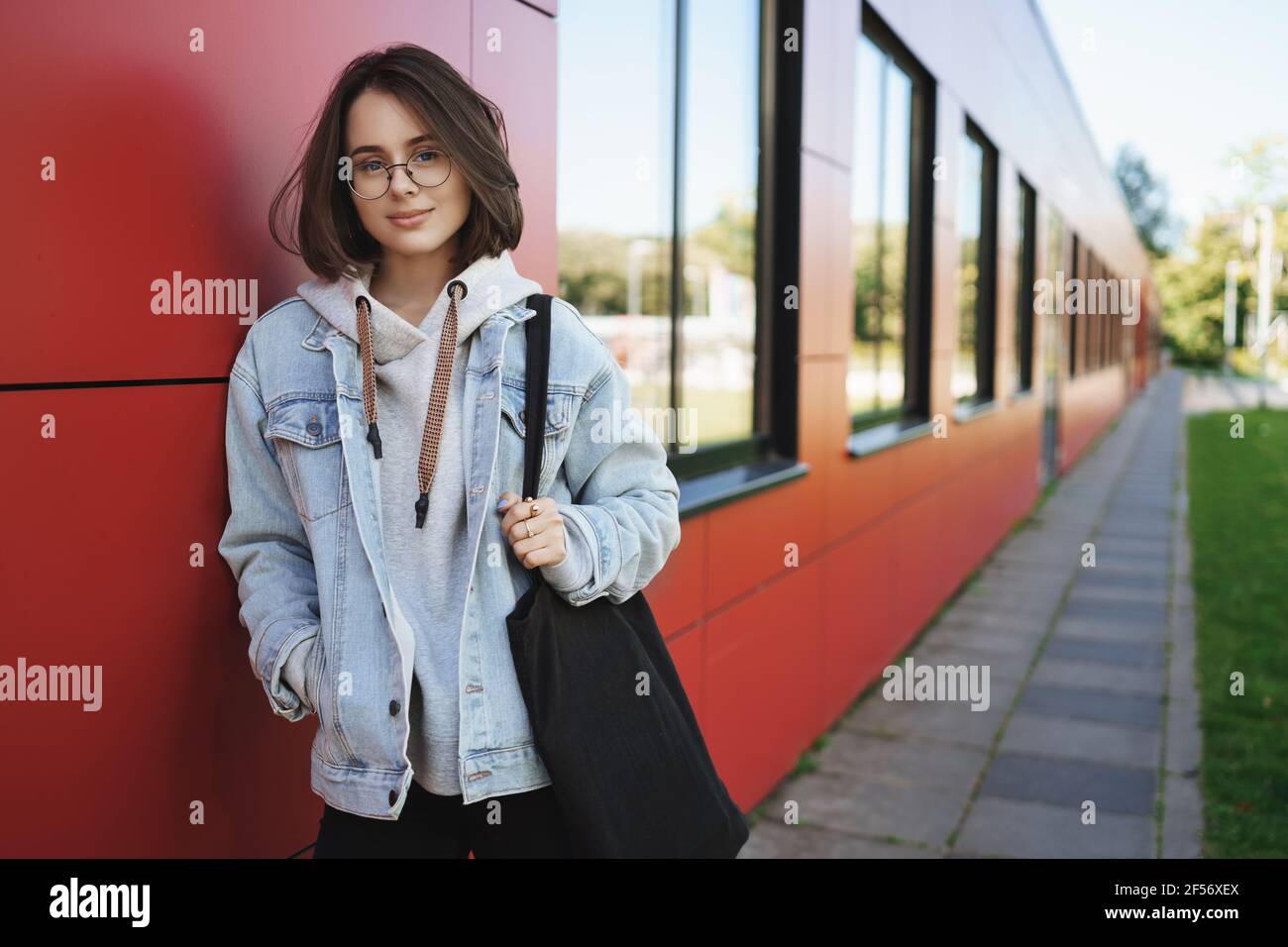Young stylish queer girl, college student leaning on wall, holding tote-bag, smiling looking at camera, walking outdoors after classes finished Stock Photo