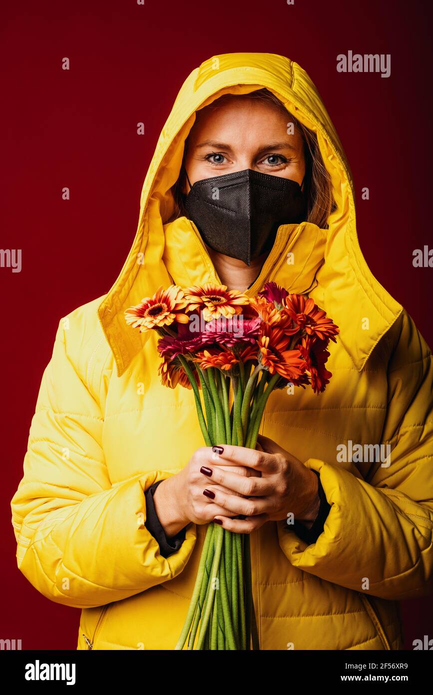 Woman wearing face mask and hooded shirt holding flower while standing against red background Stock Photo