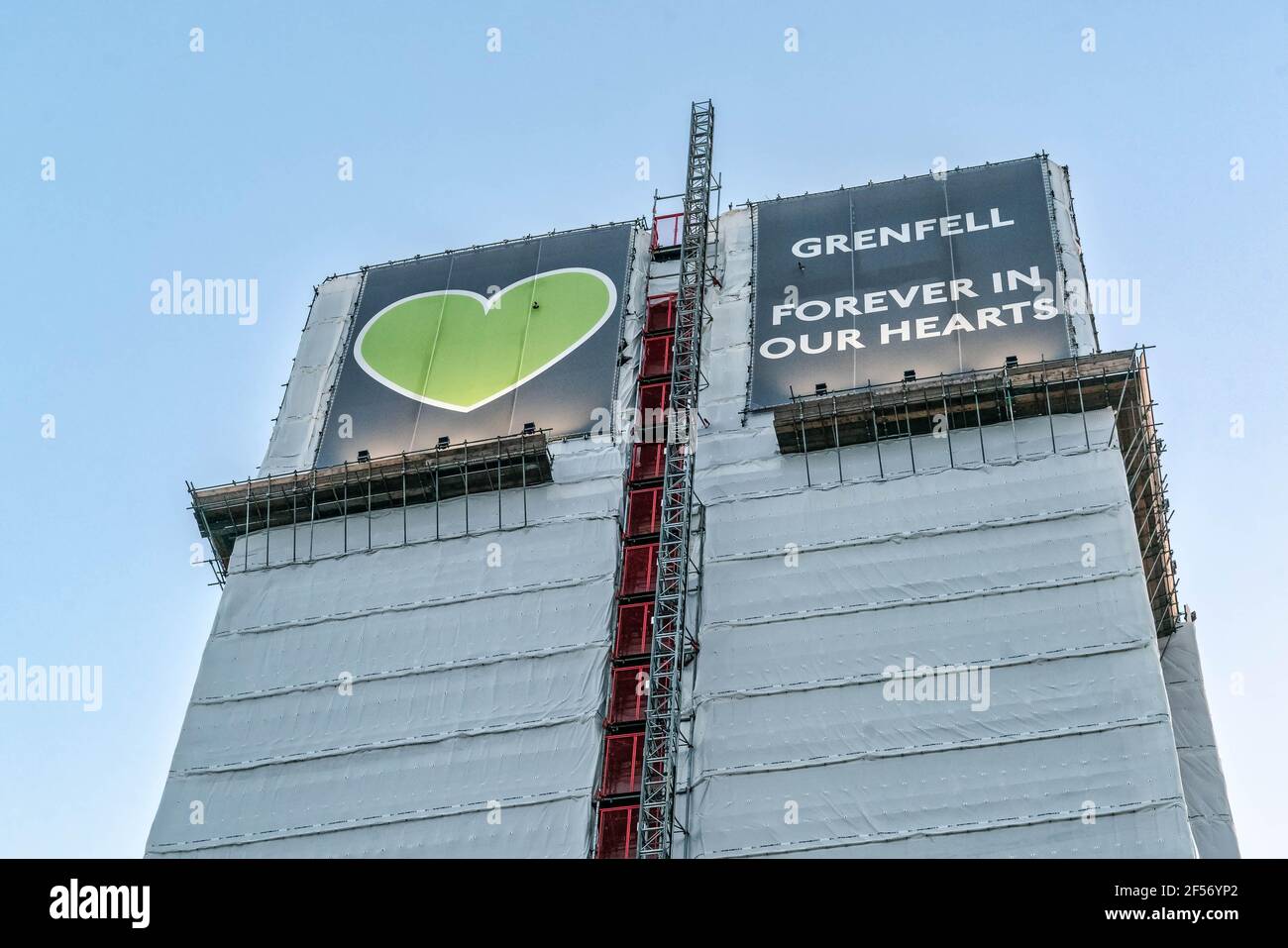 Grenfell tower cover in white tarpaulin, with the green heart logo and inscription above, stands tall against a blue sky. Stock Photo