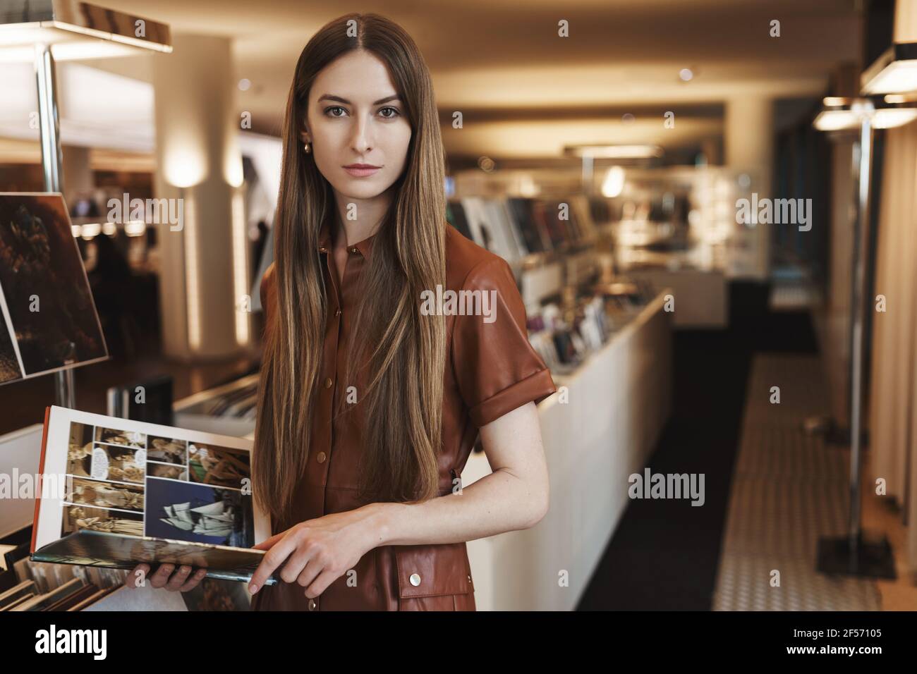 Serious-looking young female artschool student picking design magazines at vintage store, standing in stylish brown dress with fashion book, looking Stock Photo