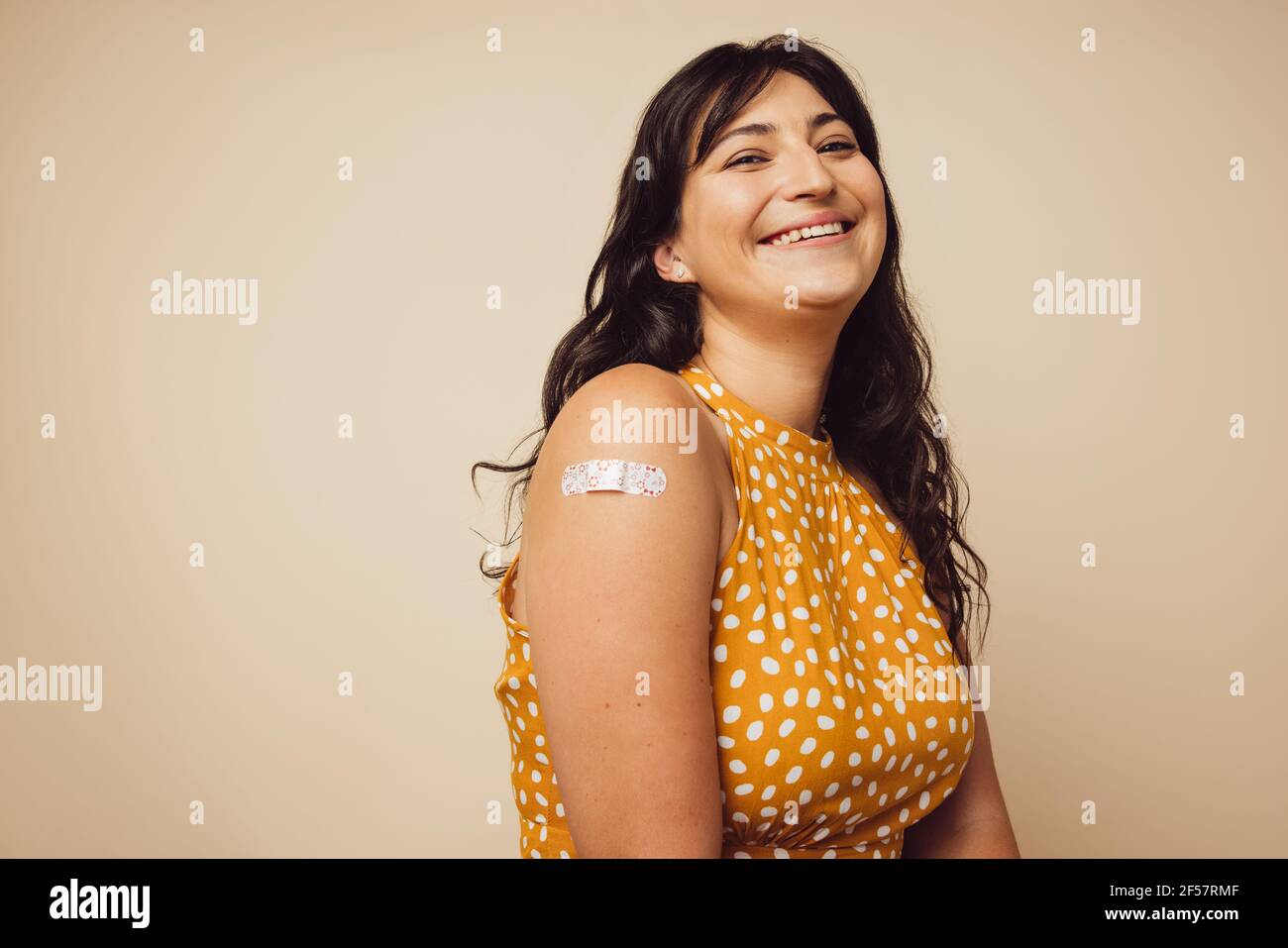 Beautiful mature with bandage on her arm after she received vaccination. Woman smiling on brown background feeling happy to get inoculated. Stock Photo
