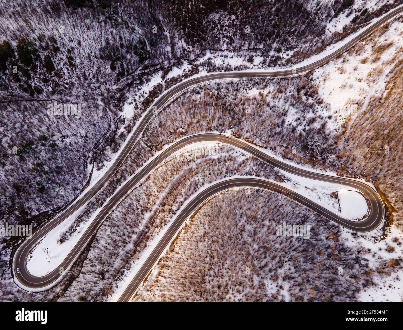 High angle aerial view drone image on the curved serpentine road trough the trees and forest in mountain range with white snow in winter day near Knja Stock Photo