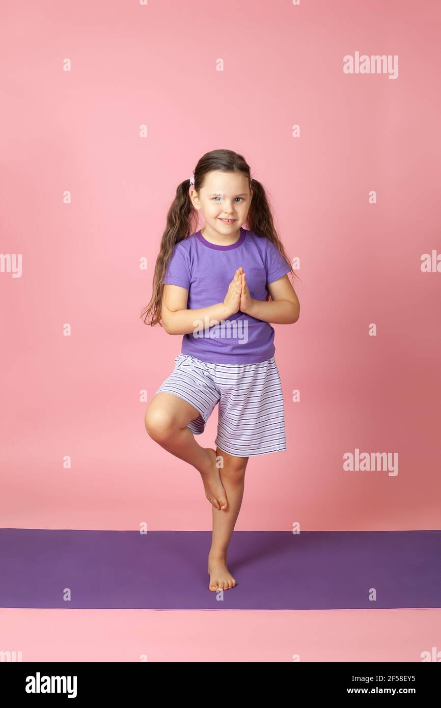 full-length portrait of a smiling girl in shorts and a T-shirt on a purple rug balancing in a tree pose in yoga, isolated on a pink background Stock Photo