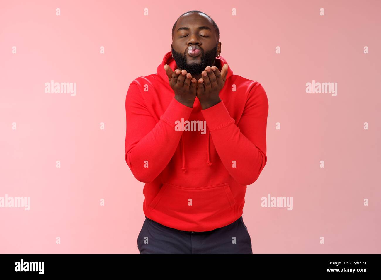 Charming cute african american bearded guy in red hoodie close eyes folding lips hold palms near mouth sending passionate air kiss blowing mwah camera Stock Photo