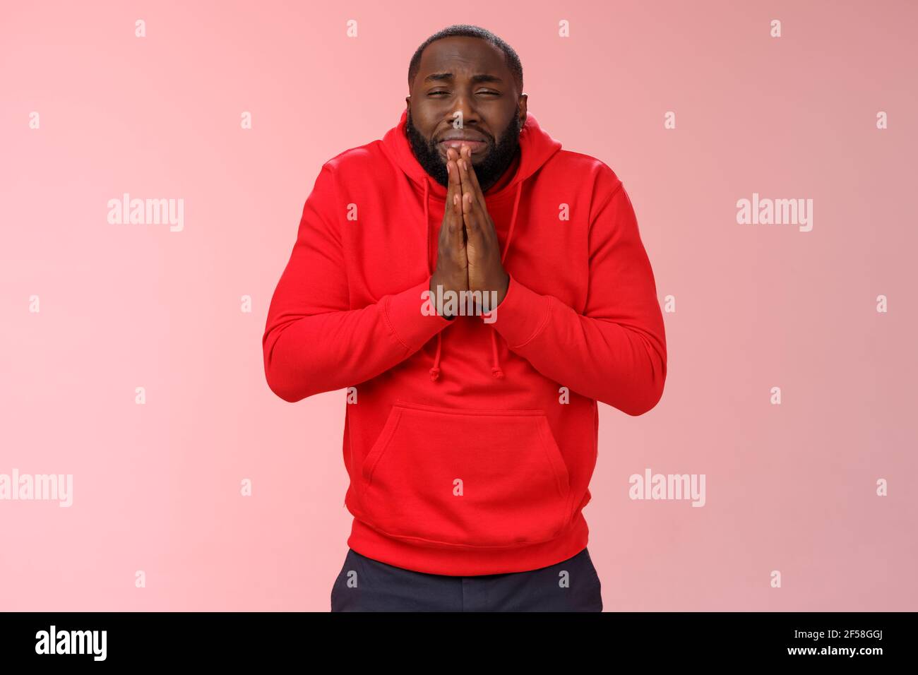 Miserable african american man beard in red hoodie stooping make supplicating upset grimace grying sobbing asking help say please hold hands pray Stock Photo