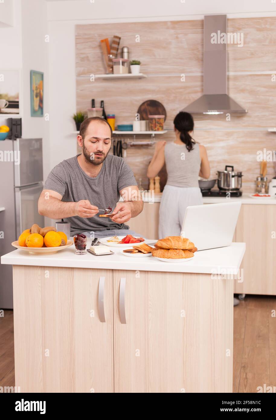Independence worker eating breakfast and working from home while his wife cooks. Freelancer working remote, talking in videoconference video call online web internet meeting from home, communication device Stock Photo