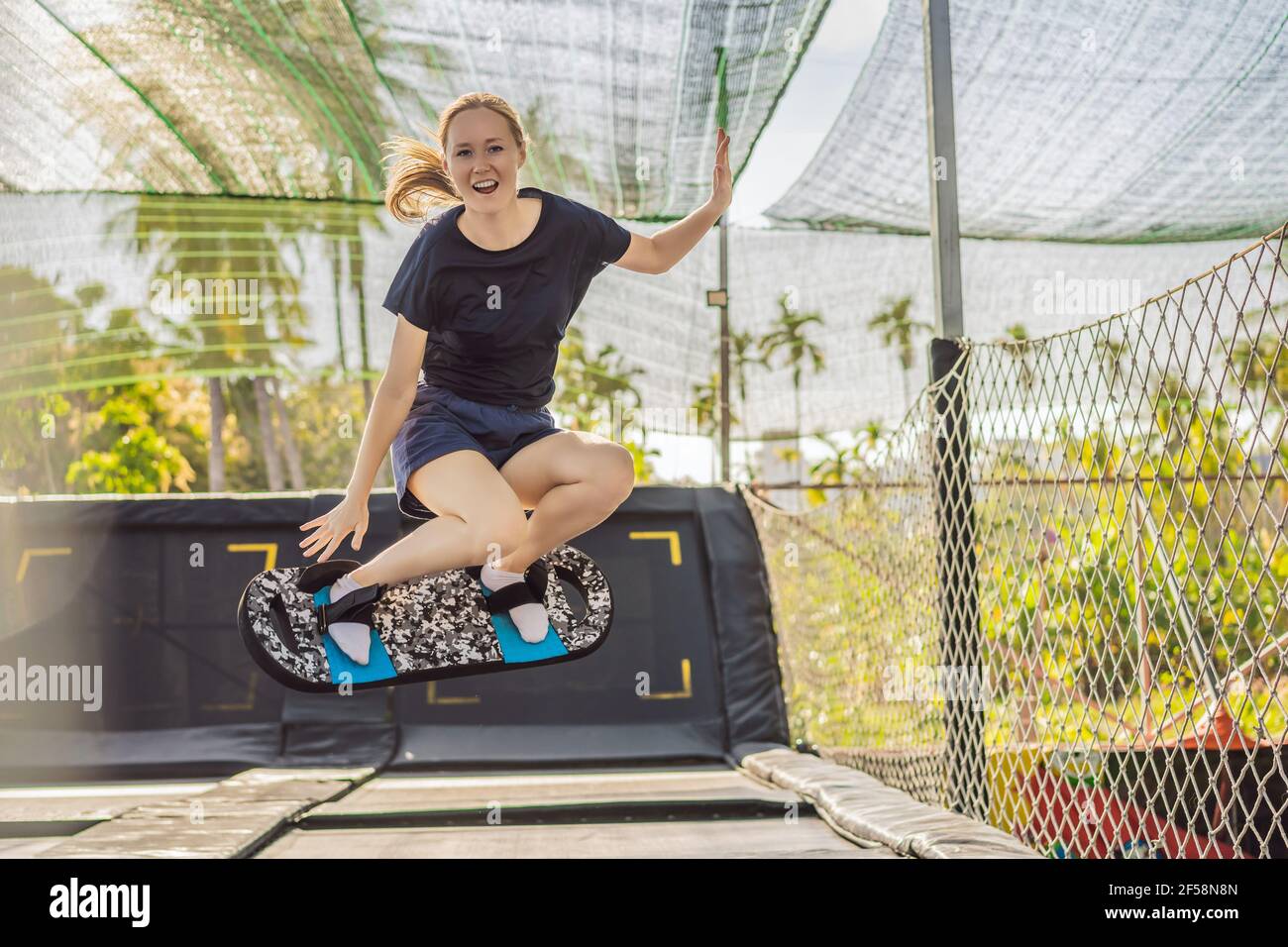 Young woman on a soft board for a trampoline jumping on an outdoor trampoline, against the backdrop of palm trees. The trampoline board is like a Stock Photo