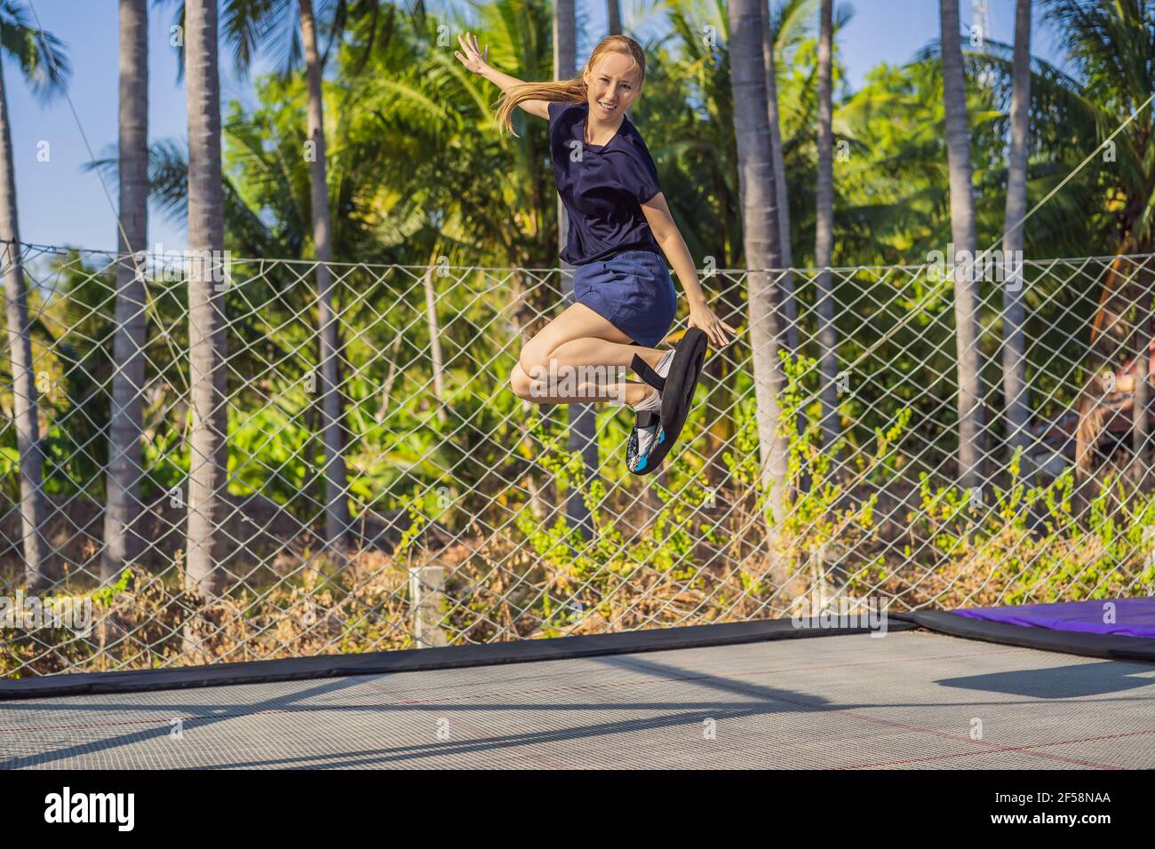 Young woman on a soft board for a trampoline jumping on an outdoor trampoline, against the backdrop of palm trees. The trampoline board is like a Stock Photo