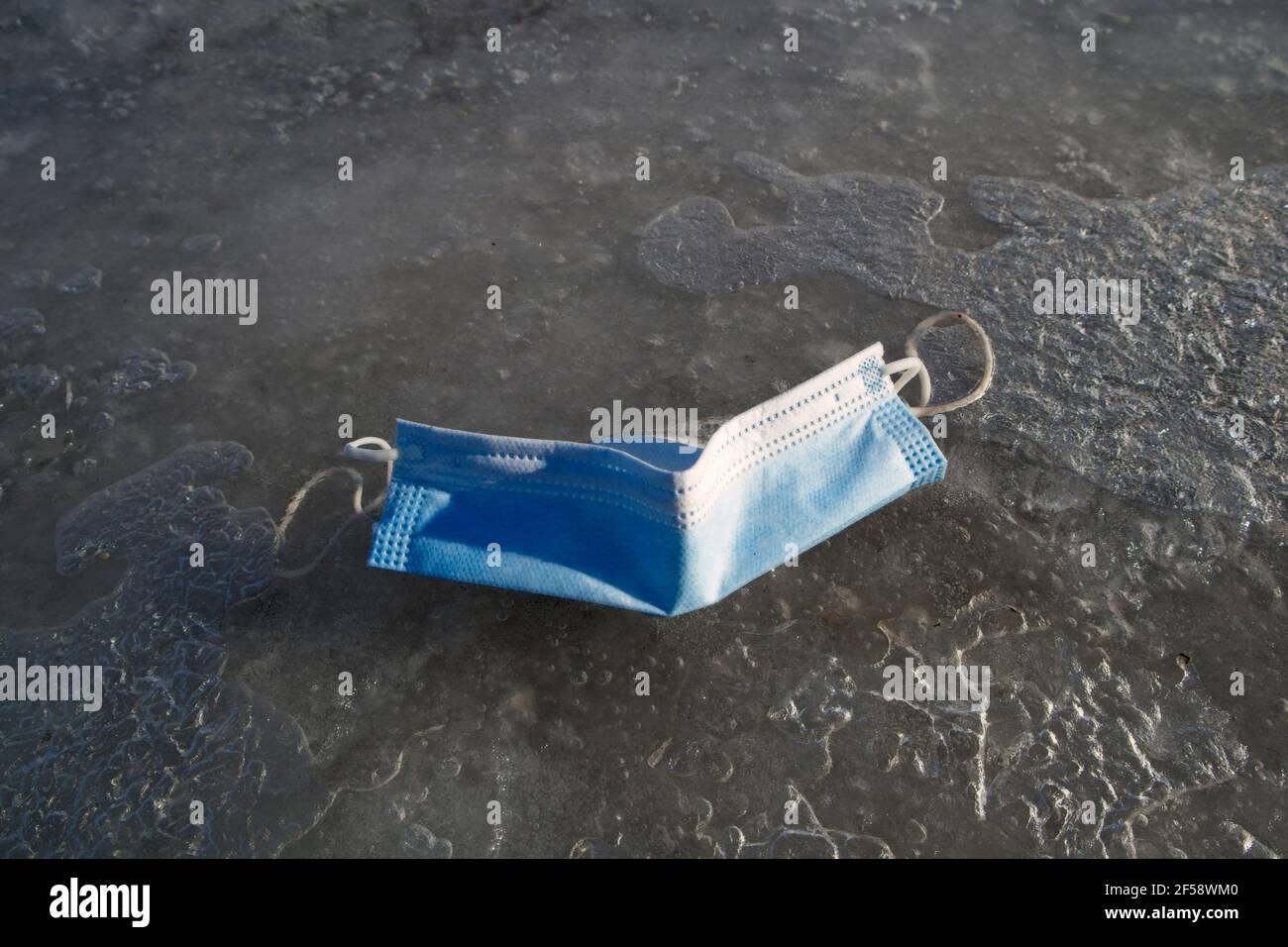 used coronavirus respiratory infection protection mask on ground in frozen puddle Stock Photo