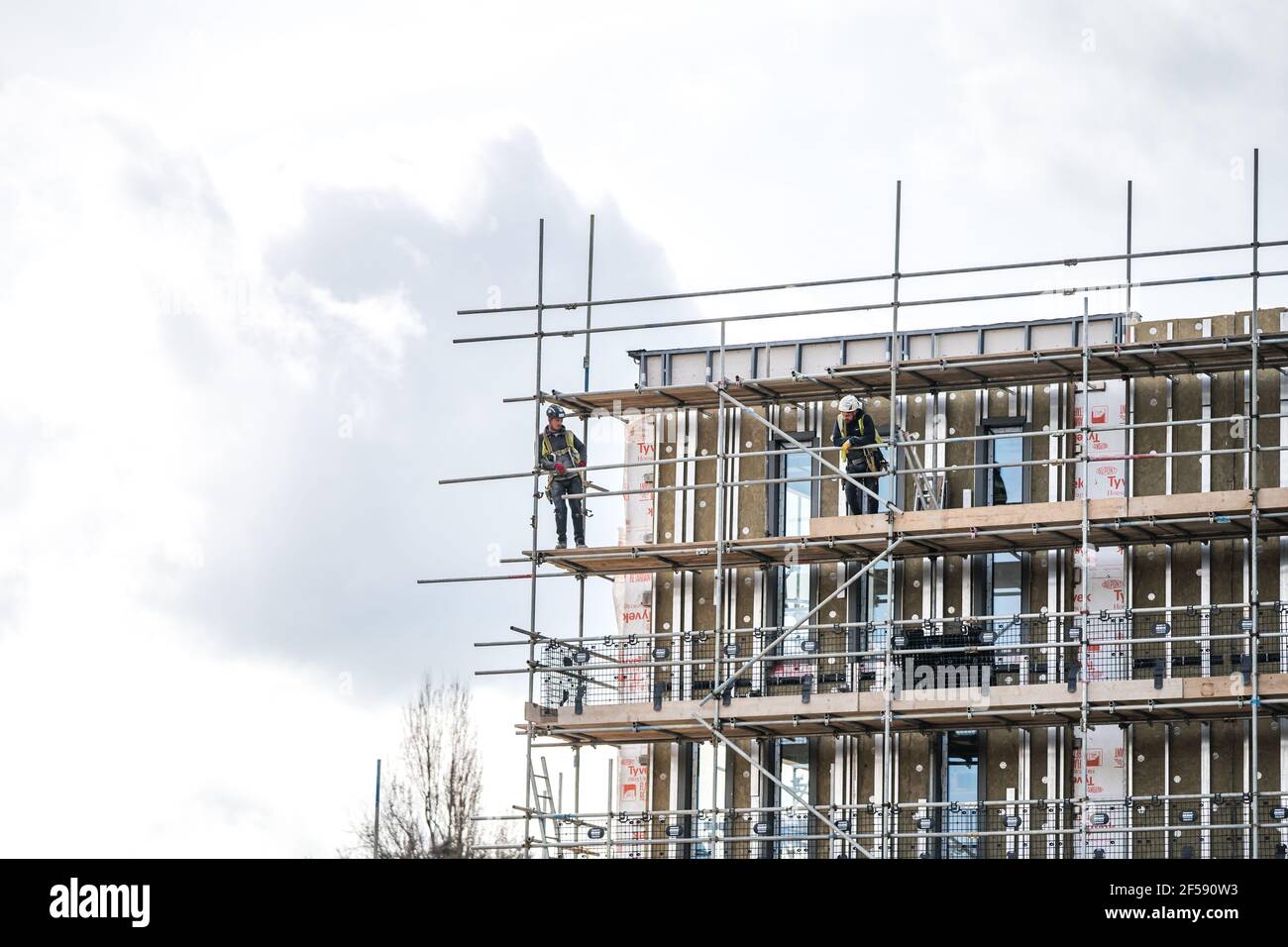 Scaffolder PPE erecting framework tall scaffolding poles high up modern new building construction site. Dangerous job safety equipment and tools Stock Photo