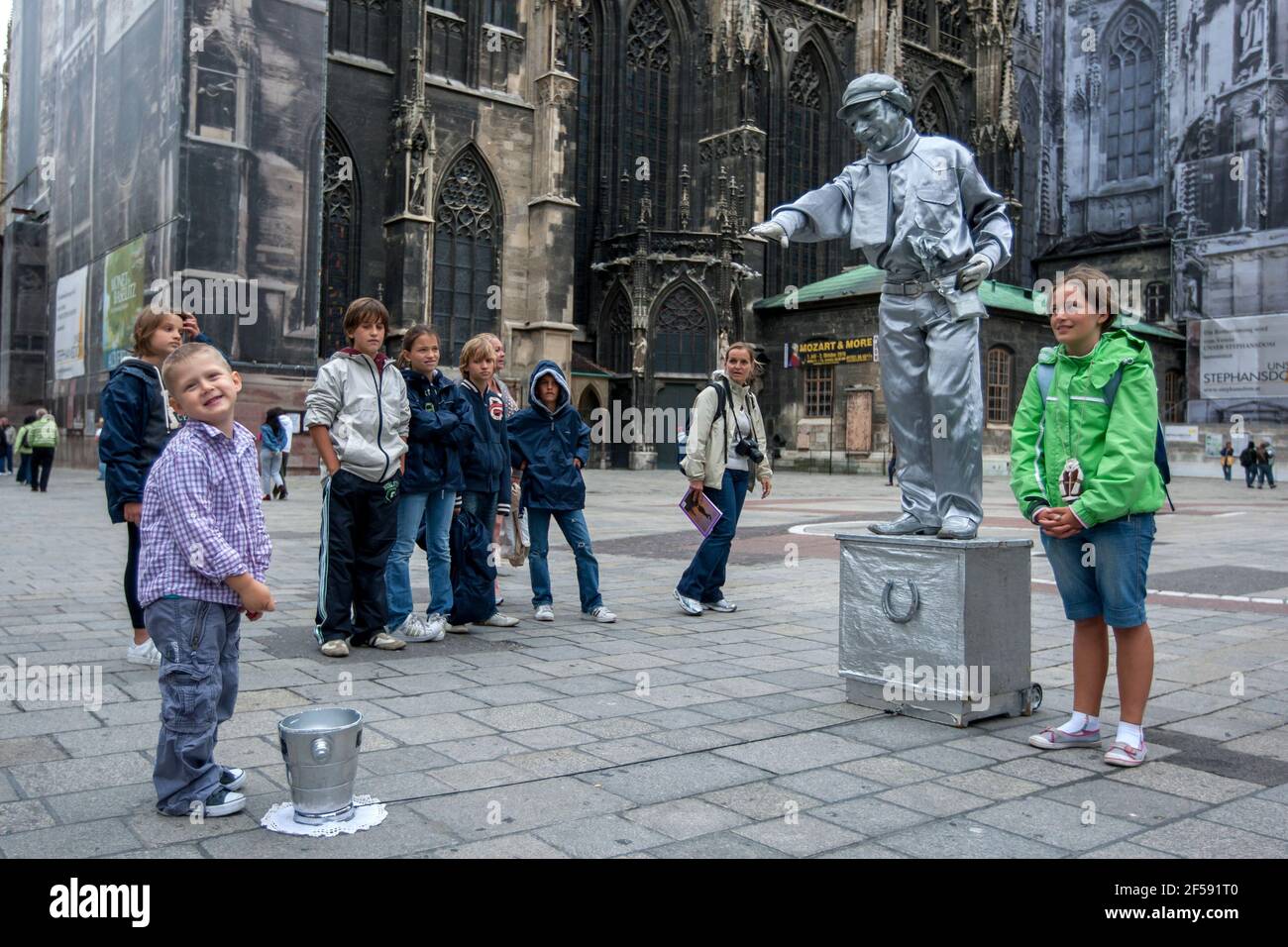 A boy drops a coin into the bucket of a statue street performer painted in silver at Vienna, Austria. In the background stands St Stephen's Cathedral. Stock Photo