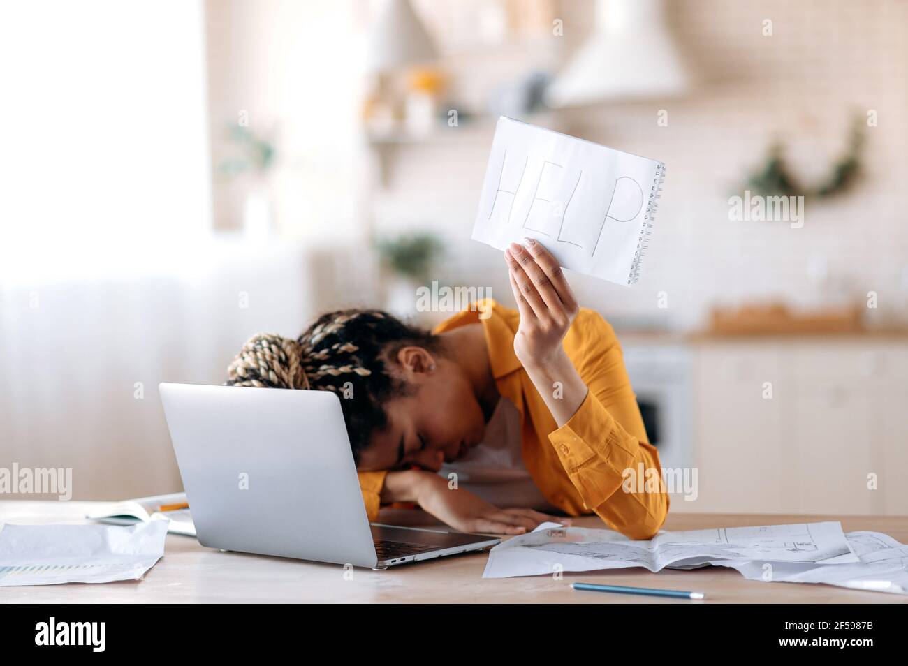 A frustrated African American female student is tired of studying at home, lies at her desk and holds a leaflet with the inscription help. Freelancer young woman need a rest from remotely work Stock Photo