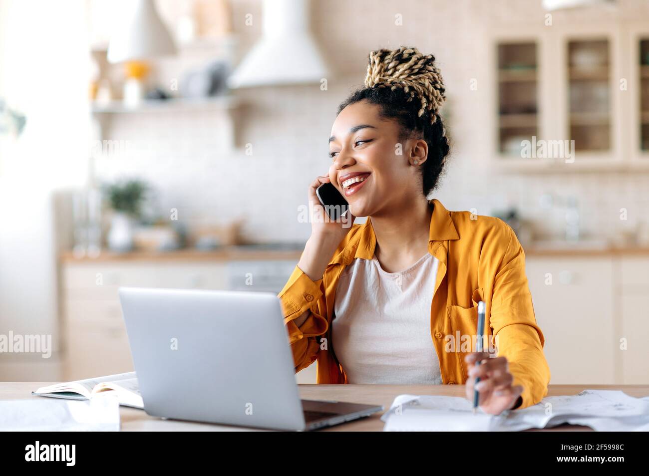 African American joyful attractive stylish young woman, freelancer, manager or real estate agent, having pleasant phone conversation with client or employee, sitting at workplace, smiling Stock Photo