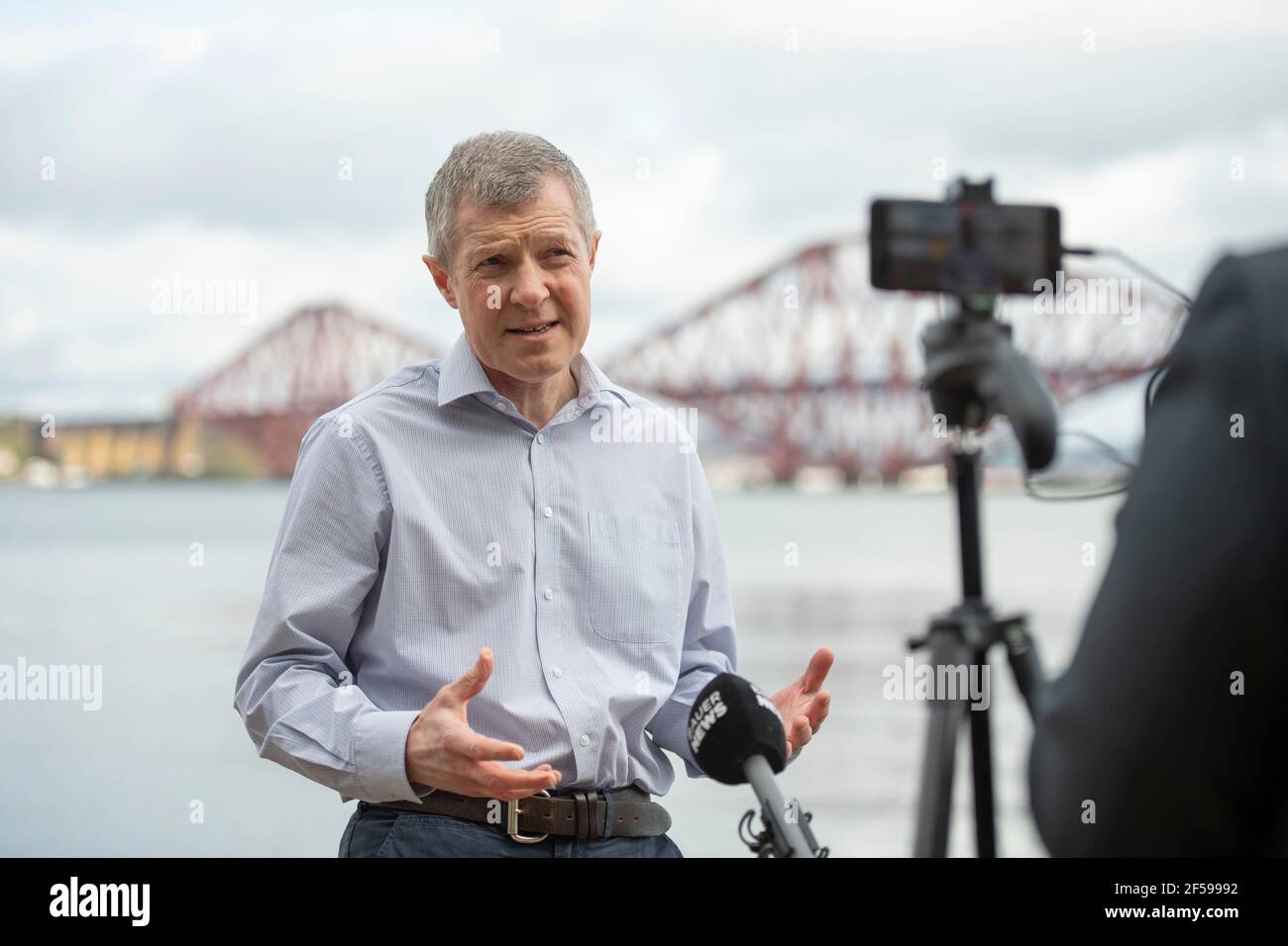 South Queensferry, Scotland, UK. 25th Mar, 2021. PICTURED: Willie Rennie MSP. Willie Rennie MSP - Leader of the Scottish Liberal Democrat Party (Scottish Lib Dems) joined by Edinburgh Northern and Leith candidate, Rebecca Bell and her daughter Daphne. He reads Rebecca's daughter a book on a giant beach chair with the view of the Forth Bridge behind as part of their Holyrood Elections campaign trail for the 6th May. Credit: Colin Fisher/Alamy Live News Stock Photo