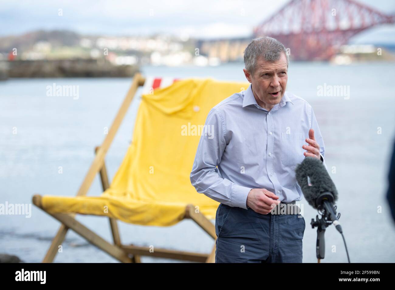 South Queensferry, Scotland, UK. 25th Mar, 2021. PICTURED: Willie Rennie MSP. Willie Rennie MSP - Leader of the Scottish Liberal Democrat Party (Scottish Lib Dems) joined by Edinburgh Northern and Leith candidate, Rebecca Bell and her daughter Daphne. He reads Rebecca's daughter a book on a giant beach chair with the view of the Forth Bridge behind as part of their Holyrood Elections campaign trail for the 6th May. Credit: Colin Fisher/Alamy Live News Stock Photo