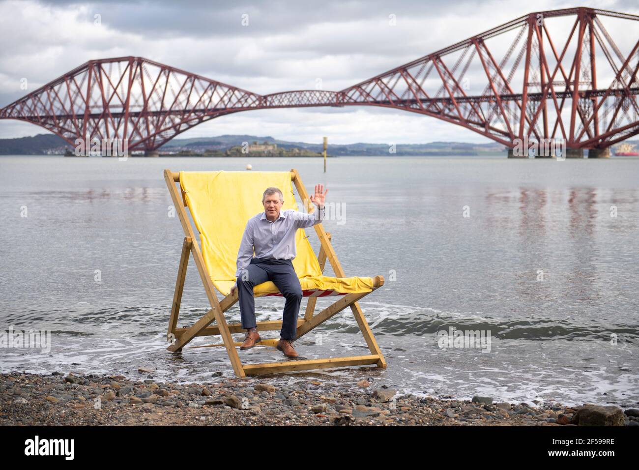 South Queensferry, Scotland, UK. 25th Mar, 2021. PICTURED: Willie Rennie MSP. Willie Rennie MSP - Leader of the Scottish Liberal Democrat Party (Scottish Lib Dems) joined by Edinburgh Northern and Leith candidate, Rebecca Bell and her daughter Daphne. He reads Rebecca's daughter a book on a giant beach chair with the view of the Forth Bridge behind as part of their Holyrood Elections campaign trail for the 6th May. Credit: Colin Fisher/Alamy Live News Stock Photo