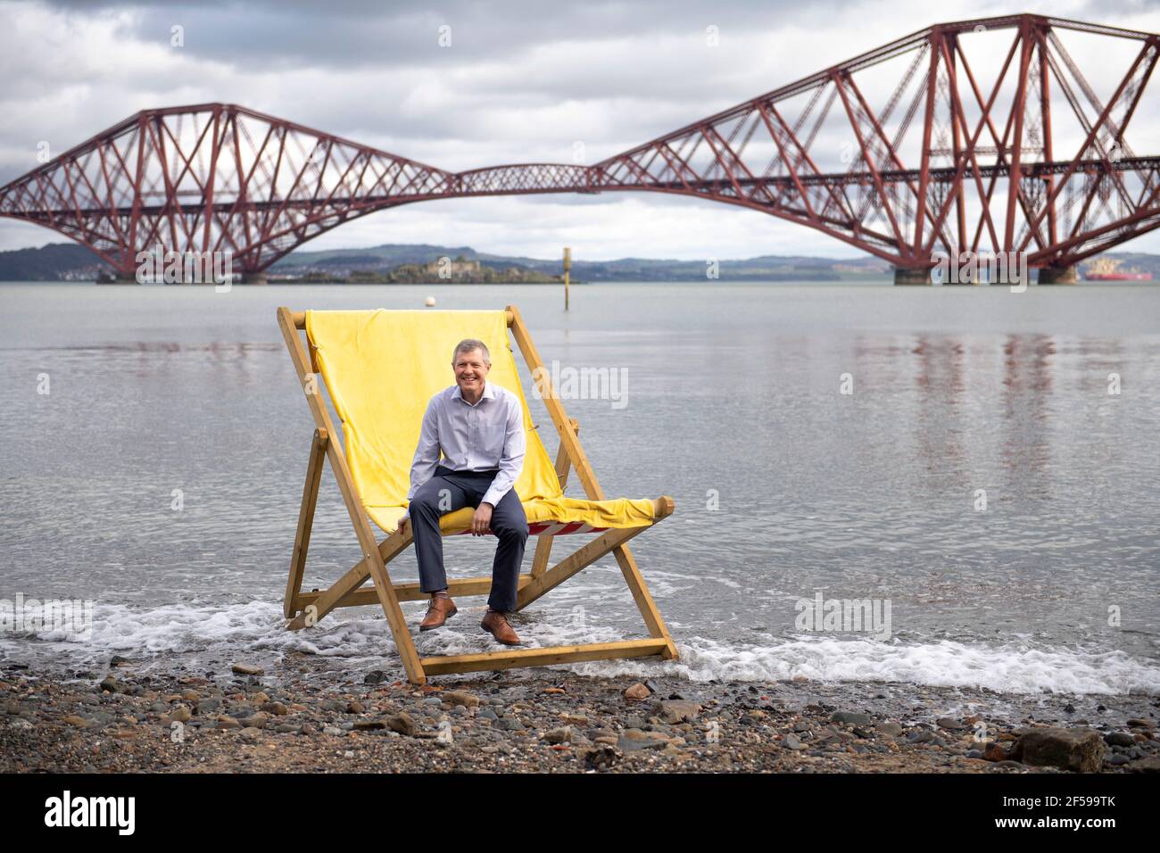 South Queensferry, Scotland, UK. 25th Mar, 2021. PICTURED: Willie Rennie MSP. Willie Rennie MSP - Leader of the Scottish Liberal Democrat Party (Scottish Lib Dems) joined by Edinburgh Northern and Leith candidate, Rebecca Bell and her daughter Daphne. He reads Rebecca's daughter a book on a giant beach chair with the view of the Forth Bridge behind as part of their Holyrood Elections campaign trail for the 6th May. Credit: Colin Fisher/Alamy Live News Stock Photo