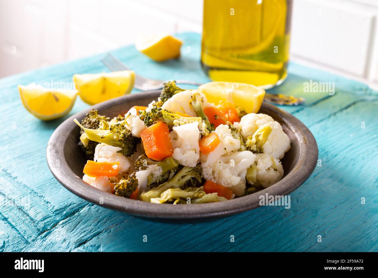 Broccoli, cauliflower and carrot salad in bowl on blue wooden table with olive oil and lemon slices. Stock Photo