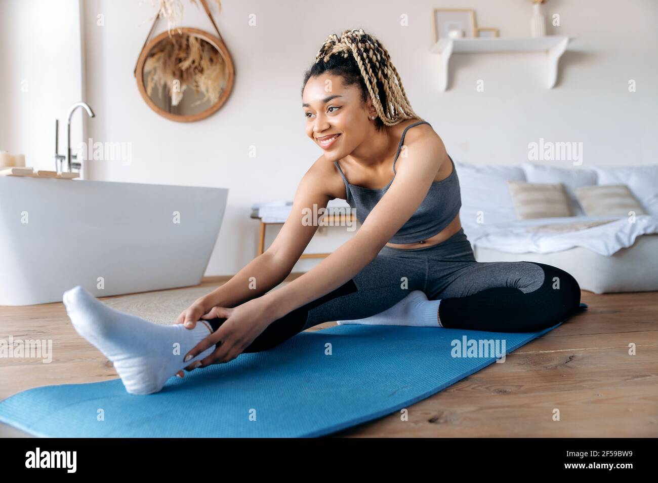 Happy joyful african american young woman in sportswear and with dreadlocks, takes care of her health, does fitness, leads a healthy lifestyle, does stretching at home on the mat, smiles Stock Photo
