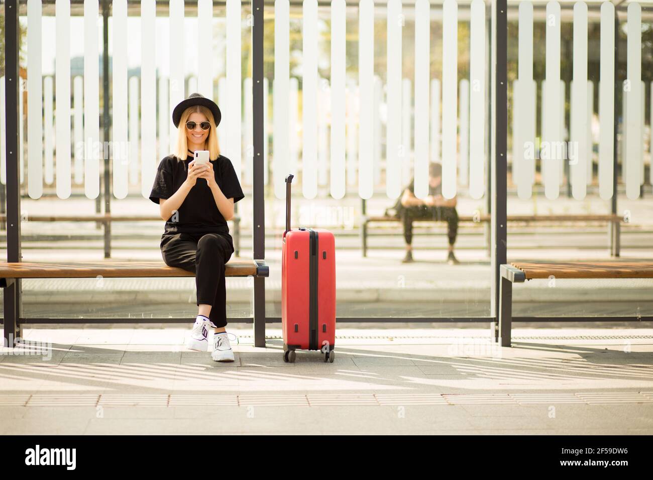 Traveler tourist woman in black casual clothes, sunglasses and hat with red suitcase use mobile phone to buy tickets online and sit on bus station. Female passenger travel abroad on weekends getaway. Air flight journey concept Stock Photo