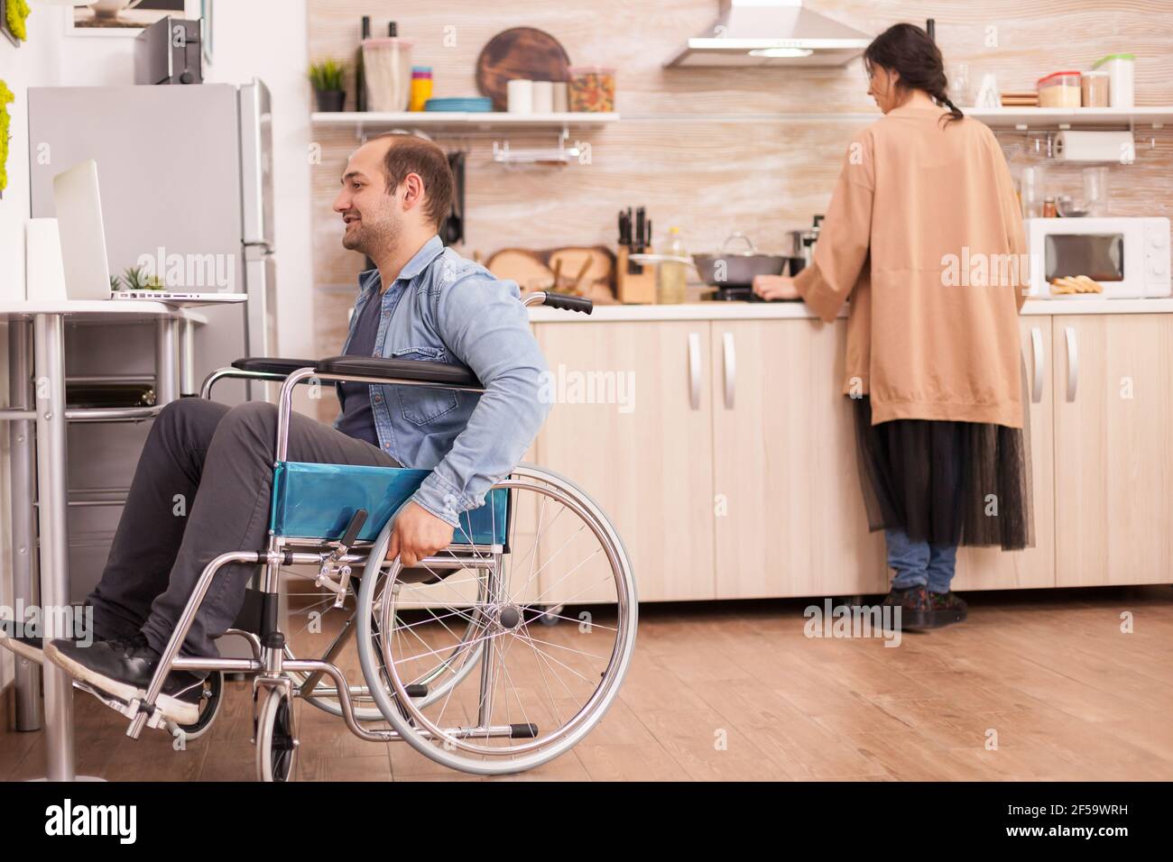 Disabled guy in wheelchair on video call in kitchen while wife cooking